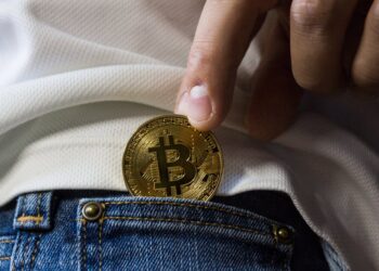 A close-up of a person's hand holding a gold-colored Bitcoin, partially tucked into the front pocket of blue jeans. The person is wearing a white shirt. The cryptocurrency is prominently displayed with a "B" and two vertical lines through it. | FintechZoom