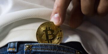 A close-up of a person's hand holding a gold-colored Bitcoin, partially tucked into the front pocket of blue jeans. The person is wearing a white shirt. The cryptocurrency is prominently displayed with a "B" and two vertical lines through it. | FintechZoom