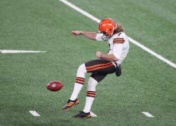 A football player in a white uniform with orange and brown accents is kicking a football on a field. He is wearing a helmet with an orange design and black cleats with orange stripes. The grass on the field is green with white yard lines marking it. | FintechZoom