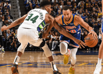 Two basketball players are in action on the court. The player on the left, wearing white with the number 34, is defending the player on the right, who is wearing a blue jersey with the number 30 and dribbling the basketball. The audience is visible in the background. | FintechZoom