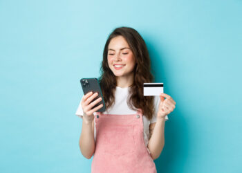 young-modern-girl-paying-online-with-credit-card-type-info-smartphone-looking-screen-with-pleased-face-shopping-app-standing-blue-background | FintechZoom
