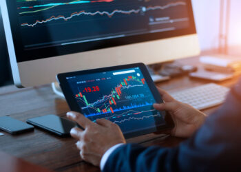 Businessman checking stock market on digital tablet and a desktop computer with stock exchange graph on screen. Financial stock market. Analyzing data in office background. | FintechZoom