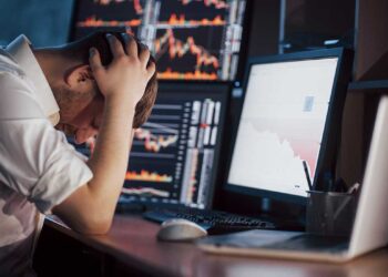 Man in desk with his hands on head near stock market screens | FintechZoom