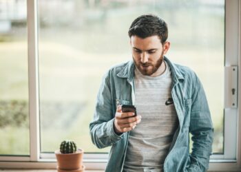 A man with a beard, dressed in a light blue denim shirt over a gray t-shirt, is leaning against a windowsill while looking at his smartphone. A small potted cactus is on the windowsill beside him, and the background shows a blurred outdoor scene—a perfect setup for checking out online casinos. | FintechZoom