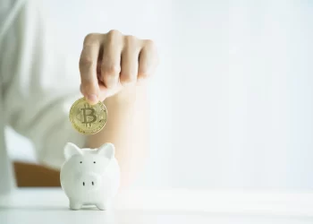 Person inserting a bitcoin on piggy bank | FintechZoom