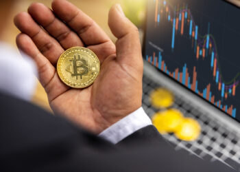 Hand holding a physical Bitcoin in hand with digital financial graphs on a laptop in the background | FintechZoom