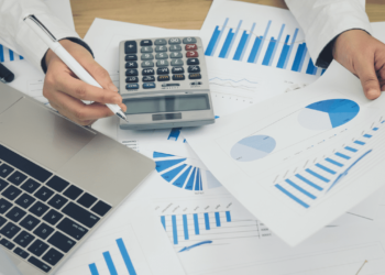 Person on desk with calculator, journal, pen, mobile phone and laptop | FintechZoom
