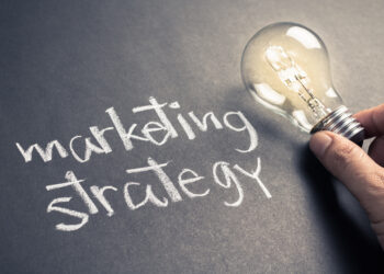 Handwriting of Marketing Strategy text on chalkboard with glowing light bulb | FintechZoom