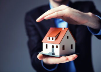 Man holding a House model in one hand and the other hand in cover the house | FintechZoom