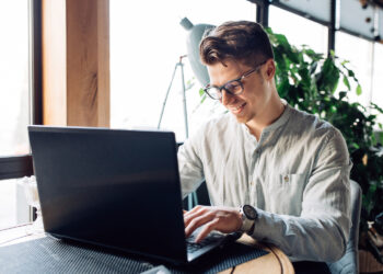 businessman in eyeglasses working on laptop, typing, spending time at cafe. | FintechZoom