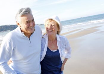 Happy senior couple walking on beach | FintechZoom