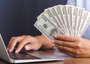 Person using laptop and holding 100 USD notes | FintechZoom