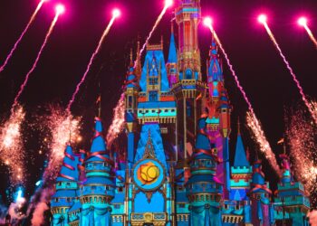 A colorful castle is illuminated at night with vibrant lights and projections. Fireworks explode above, and smoke can be seen around the castle. Silhouetted people, possibly including a Pepperboy vendor, are gathered below, watching the dazzling display. | FintechZoom