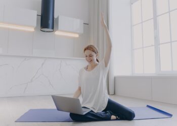 Young woman sitting on her knees on floor with laptop online fitness | FintechZoom