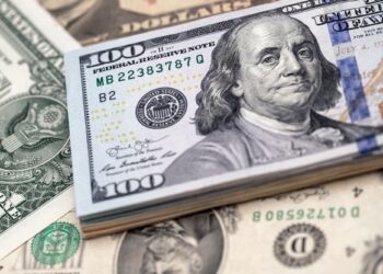 A stack of U.S. one-hundred-dollar bills featuring Benjamin Franklin is placed on top of various other U.S. currency, including a one-dollar bill with George Washington. The arrangement highlights the different denominations and designs of the currency. | FintechZoom