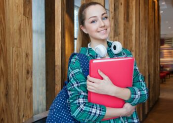 Student Standing in Hallway While Holding Book | FintechZoom