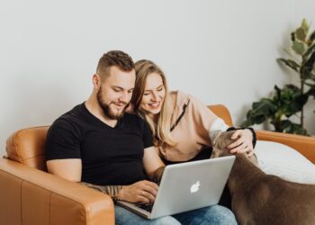 Happy couple in sofa with their dog using laptop | FintechZoom