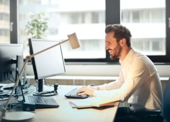 Man in White Dress Shirt Sitting on Black Rolling Chair While Facing Black Computer Set and Smiling | FintechZoom
