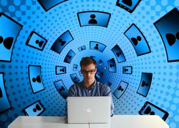 A person with glasses sits at a table using a laptop. The background features a blue, tunnel-like pattern with multiple black silhouettes on screens, creating an illusion of layers of digital connections, suggesting heavy online engagement—perhaps even using iganony to navigate through social networks discreetly. | FintechZoom