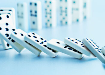 Close-up of white dominoes falling in a curved line on a light blue surface, reminiscent of crypto falling in value, with more blurred dominoes standing in the background. The dominoes have black dots indicating their respective values. The image captures the motion and continuity of the falling dominoes. | FintechZoom
