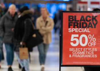 A promotional sign in a busy mall advertises Black Friday Savings, offering 50% off select styles of cosmetics and fragrances. Shoppers walk by the sign, carrying bags and engaging in shopping activities. | FintechZoom