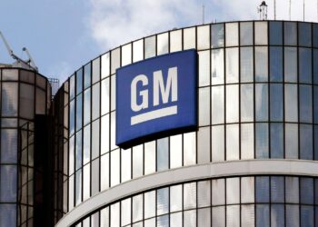The image shows the exterior of a tall, glass building with the General Motors (GM) logo prominently displayed on a blue sign at the top. The sign features the white GM letters on a blue background, with a white underscore below the letters—an iconic symbol for GM stock investors. | FintechZoom