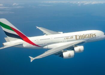 An Emirates Airbus A380 is in mid-flight above a vast expanse of blue ocean. The airplane's white body features the airline's logo and green, white, red, and black tail design. Amidst a clear sky with scattered clouds, this scene reflects a promising market outlook for international travel. | FintechZoom