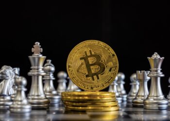 A gold Bitcoin coin stands upright surrounded by silver chess pieces, invoking a scene straight out of Crypto Chess. Stacked Bitcoin coins lie below, all illuminated against a dark backdrop that accentuates the focal elements. | FintechZoom