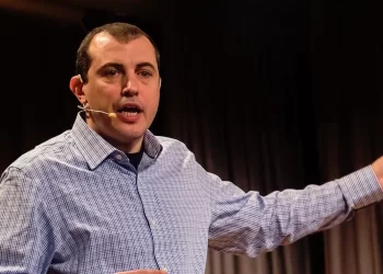 A man wearing a checked shirt and a headset microphone is speaking passionately about crypto influencers and pointing to his right against a backdrop of dark curtains. | FintechZoom