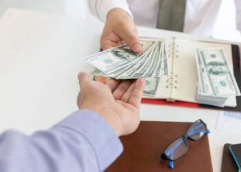 One person hands a stack of U.S. dollar bills to another over a desk, signaling they’ve been approved for a loan. On the desk, there's a notebook with more money inside it, eyeglasses, and a brown folder. Both individuals are dressed in business attire. | FintechZoom