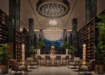 Luxurious hotel lobby featuring a grand chandelier, marble columns, and a central bar with high-backed stools. Despite its opulent look, budget-conscious travelers will be pleased to find elegant seating, adorned plants, wooden shelves, and tall racks that create an inviting and sophisticated atmosphere. | FintechZoom