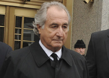An older man with gray hair and a serious expression, resembling Bernie Madoff, is wearing a black overcoat and white shirt with a black tie. He stands in front of a building with golden doors and glass panels, while another person in black clothing appears blurry in the background. | FintechZoom