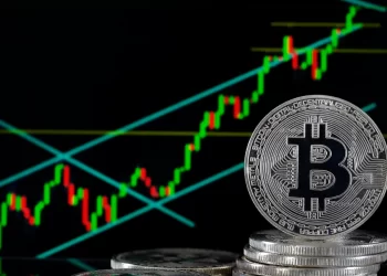 A stack of physical Bitcoin coins is shown in front of a screen displaying a rising cryptocurrency price chart. The green and red candlesticks on the chart indicate upward market trends, with blue trend lines marking the price's movement, aligning with optimistic Bitcoin price predictions. | FintechZoom