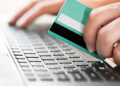 A person's hand holding a debit or credit card above a laptop keyboard suggests an online payment or purchase. The other hand is typing on the keyboard, indicating ease of use. The green card with a white and black stripe signifies convenience, perhaps even to buy Bitcoin with your credit card. | FintechZoom
