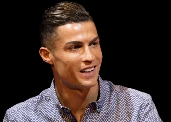 A person with short, slicked-back hair is smiling and looking slightly to their right, reminiscent of Cristiano Ronaldo. They are wearing a collared, patterned shirt with a dark, abstract design against a black background. | FintechZoom