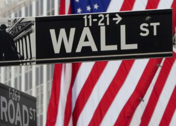 Street signs for Wall Street and Broad Street at an intersection in New York City, where discussions about the most popular stocks often occur. An American flag prominently hangs in the background, partially visible on the right side. The Wall Street sign features an arrow and the numbers 11-21 with a graphic of a statue. | FintechZoom