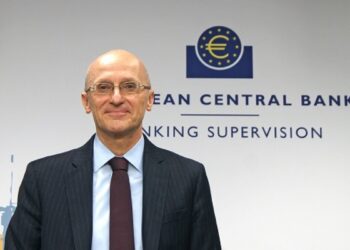A man in a suit and tie stands smiling in front of a wall with the text "EUROPEAN CENTRAL BANK BANKING SUPERVISION" and the European Central Bank logo, which features a stylized euro symbol inside a circle of stars, reflecting the intersection of traditional banking and emerging trends like Crypto News. | FintechZoom