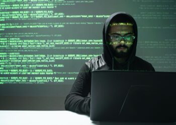 A person wearing a hoodie and glasses is typing on a laptop in a dimly lit room. Lines of green code are projected on the wall behind them, creating a focused and intense atmosphere. The coding reflects the person's involvement in programming or fraud detection work. | FintechZoom