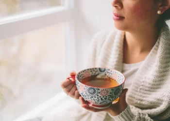 A person wearing a cream-colored knitted sweater holds a large, ornate floral-patterned mug filled with cannabis tea while sitting near a bright window. The person is looking out the window, creating a calm and reflective atmosphere. | FintechZoom