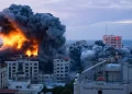 A cityscape is shrouded in thick, dark smoke and flames, with a large building on fire amidst a densely packed area of high-rise structures. The sky is clouded, and there are signs of destruction and damage to nearby buildings that began despite the four-day ceasefire on Markets. | FintechZoom