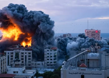 A cityscape is shrouded in thick, dark smoke and flames, with a large building on fire amidst a densely packed area of high-rise structures. The sky is clouded, and there are signs of destruction and damage to nearby buildings that began despite the four-day ceasefire on Markets. | FintechZoom