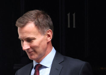 A man in a suit and tie, with short brown hair, is looking downwards with a serious expression. The door behind him is black and has the number 11 on it, suggesting he might be formulating an important Autumn Statement. | FintechZoom