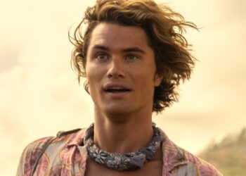A person with wavy hair and a patterned shirt, reminiscent of John B from Outer Banks, stares intently ahead. They wear a bandana around their neck, and the background features a cloudy sky with hints of vegetation. The warm lighting gives the scene a golden glow. | FintechZoom