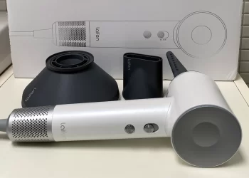 A white Laifen hair dryer with a sleek design is displayed on a cream-colored surface. The dryer has two buttons and a circular air intake. Next to it are three black attachments. The packaging box in the background shows a diagram of the product and the Lalfin brand name. | FintechZoom