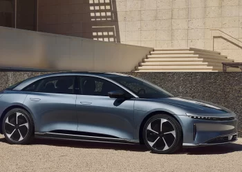 A sleek, metallic silver electric sedan is parked in front of a modern building with tall, beige stone walls and a staircase in the background. The car features a low profile, black roof, and futuristic design accents, including thin LED headlights—a testament to the adoption of electric vehicles. | FintechZoom