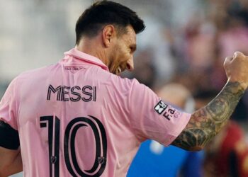 A soccer player wearing a pink jersey with the name "Messi" and the number 10 celebrates a goal with a fintech-inspired fist pump. The player has tattoos on their right arm and is wearing a black captain's armband. The background shows a blurred stadium and spectators. | FintechZoom