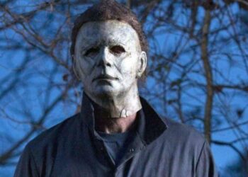 A person is wearing a weathered, white mask with dark, hollow eyes and unkempt hair. Dressed in a dark jumpsuit reminiscent of Michael Myers, they stand outdoors with leafless branches visible in the background. The scene has an eerie, unsettling atmosphere. | FintechZoom