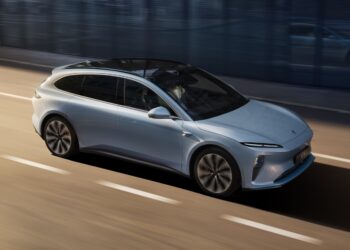 A sleek, silver Tesla electric vehicle with a glossy finish is driving on a smoothly paved road. The car features a panoramic sunroof and aerodynamic design. The background shows a blurred cityscape, indicating the car is in motion. | FintechZoom
