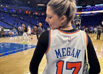 A woman with her hair in a bun stands on a basketball court, wearing a white jersey with "Overtime Megan" and the number "17" on the back. People are seen in the background, and the arena is filled with blue and orange colors. | FintechZoom
