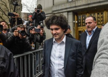 A man with curly hair and wearing a dark suit walks through a crowd of photographers and reporters. He appears to be escorted by security personnel, perhaps aware that the bigger they come, the harder they fall. The background features a large, ornate, gold-colored doorway. | FintechZoom